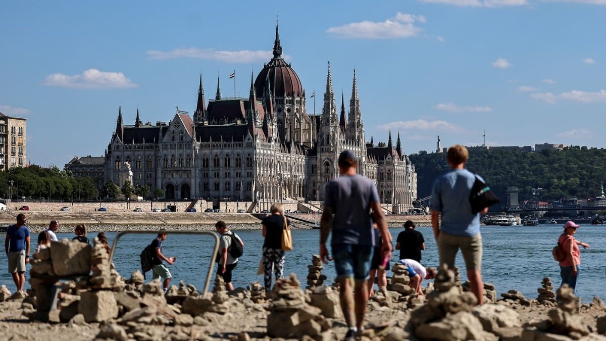 albia secată a Dunării în Budapesta, oferind o perspectivă inedită asupra orașului. În prim-plan și în planul median se întinde o suprafață vastă și accidentată, acoperită cu numeroase pietre de dimensiuni variate, bolovani și resturi de rocă, indicând un nivel extrem de scăzut al apei. Pe această întindere expusă, vizitatorii au construit o multitudine de mici turnulețe din pietre suprapuse, creând un peisaj texturat. Mai multe persoane sunt surprinse plimbându-se direct pe albia uscată, unii stând în picioare sau fotografiind împrejurimile cu telefoanele mobile. Dincolo de această zonă pietroasă și de o porțiune mult îngustată a fluviului, pe malul opus, domină peisajul arhitectural clădirea monumentală a Parlamentului maghiar, o structură grandioasă caracterizată printr-o cupolă centrală proeminentă și zeci de turnuri ascuțite. În stânga acesteia se înșiruie alte clădiri urbane masive cu mai multe etaje, așezate de-a lungul falezei presărate cu rânduri de copaci.