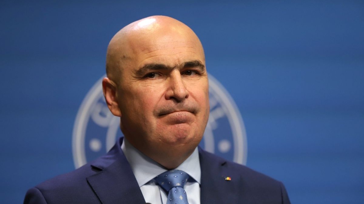 Premierul Ilie Bolojan, cu costum și cravată, îți mușcă buzele