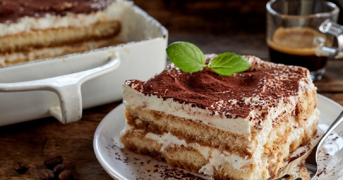 Rețete de tiramisu - Tiramisu clasic, așezat pe o farfurie, cu cacao deasupra și o ceașcă de cafea în fundal