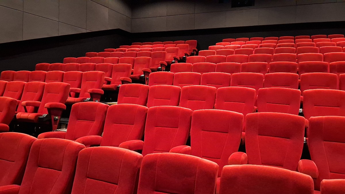Imaginea prezintă interiorul unei săli de cinema moderne, cu rânduri ordonate de scaune roșii intens luminate.
