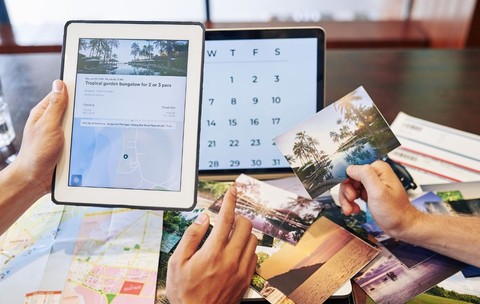 O imagine filmată de sus arată niște mâini care țin o tabletă pe ecranul căreia apare un bungalou tropical, precum și un calendar afișat pe un laptop