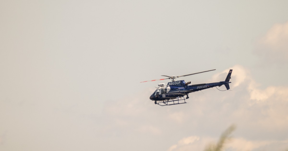 Imaginea surprinde un elicopter de culoare închisă (albastru sau negru) în zbor, pe un fundal de cer palid, ușor înnorat. Pe corpul aeronavei se distinge clar codul de înregistrare N803CP și textul "POLICE" (Poliție), indicând că este vorba despre un elicopter de supraveghere sau intervenție.