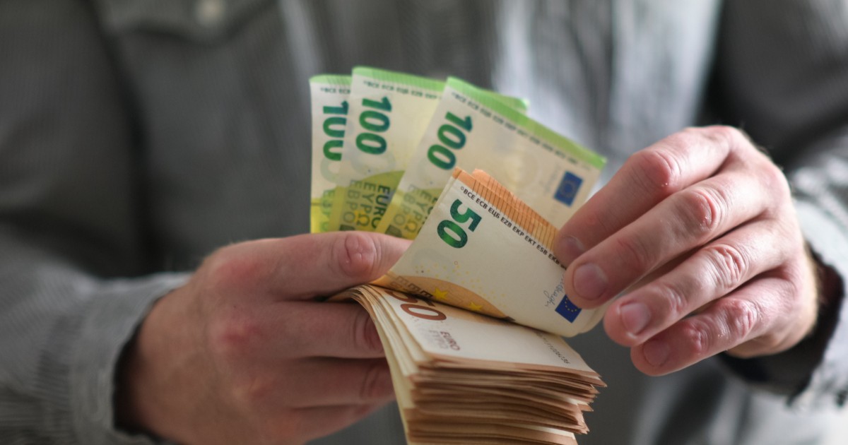 Un bărbat pensionar cu cămașă gri care ține în mână mai multe hârtii de 50 și 100 de euro