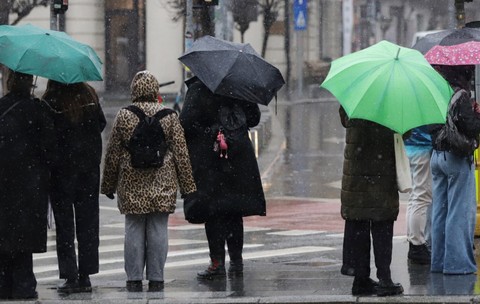 Mai mulți pietoni așteptând la o trecere de pietoni pe o vreme ploioasă și rece, adăpostindu-se sub umbrele colorate și purtând haine groase.