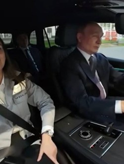 Vladimir Putin conduce un SUV Volga, îmbrăcat într-un costum închis la culoare și cravată. Pe scaunul din dreapta se află o femeie purtând centura de siguranță, iar în spate se vede un alt pasager. Imaginea este surprinsă din interiorul mașinii, în timp ce vehiculul se deplasează printr-o zonă industrială.