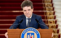Nicușor Dan îmbrăcat în costum bleumarin și cravată cu model, privind spre cameră din spatele unui pupitru oficial.