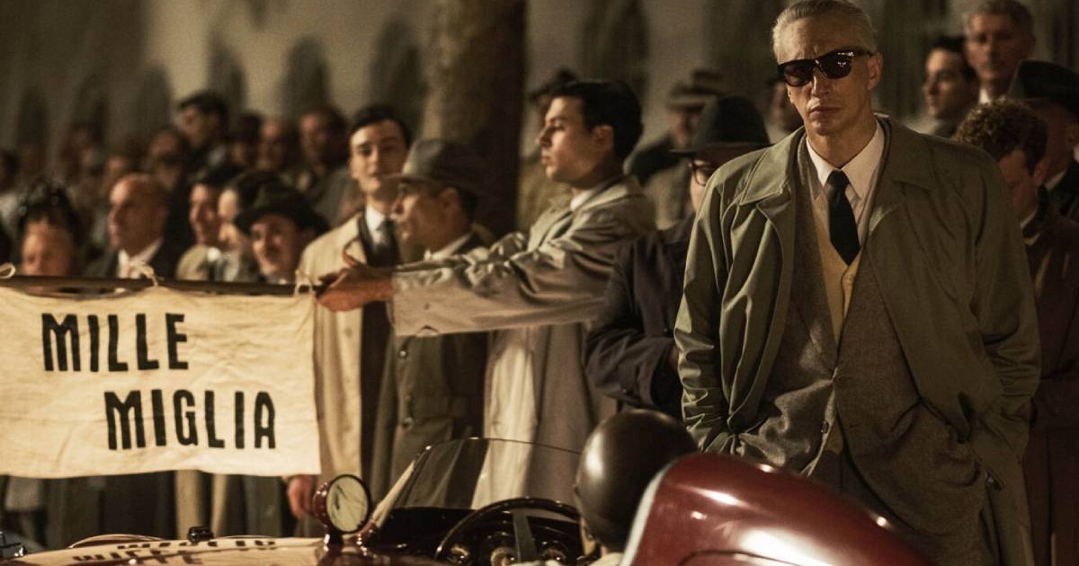 Scena din filmul FERRARI, 2023, se vede Adam Driver in prim plan, în rolul Enzo Ferrari, in spatele unei masini Ferrari rosie, in spate o multime de oameni, figuranti