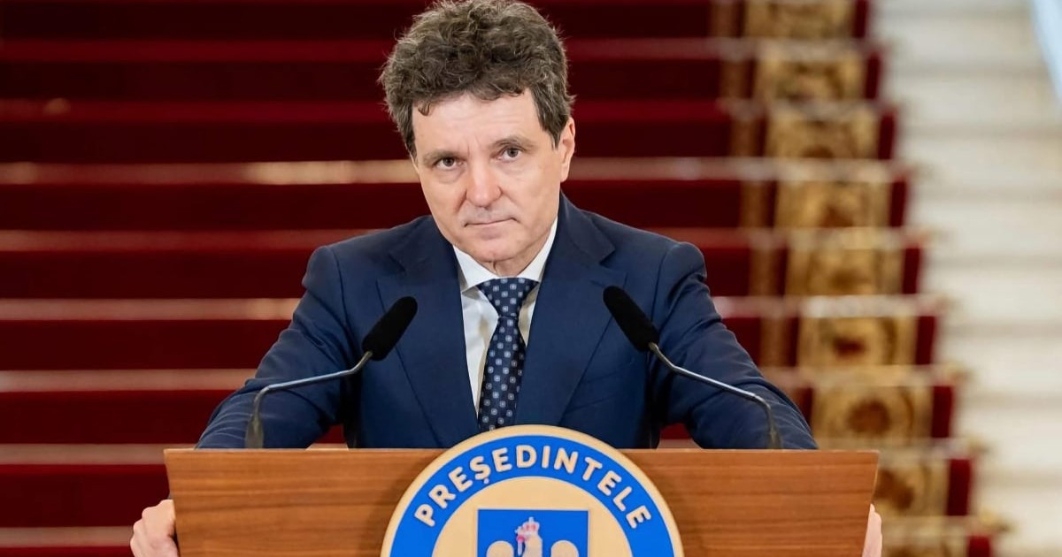 Nicușor Dan îmbrăcat în costum bleumarin și cravată cu model, privind spre cameră din spatele unui pupitru oficial.
