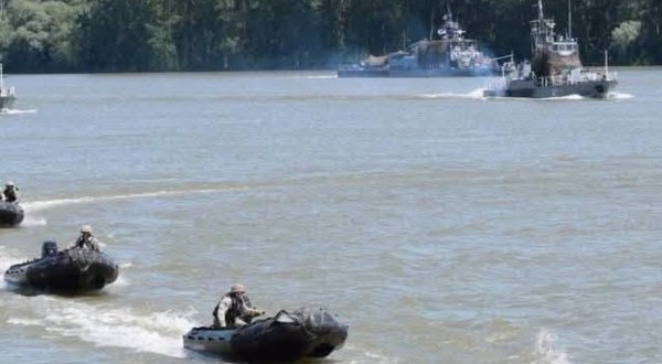 Un exercițiu militar pe apă, în care mai multe bărci pneumatice rapide ghidate de militari înaintează pe un fluviu, având nave militare de patrulare în fundal.