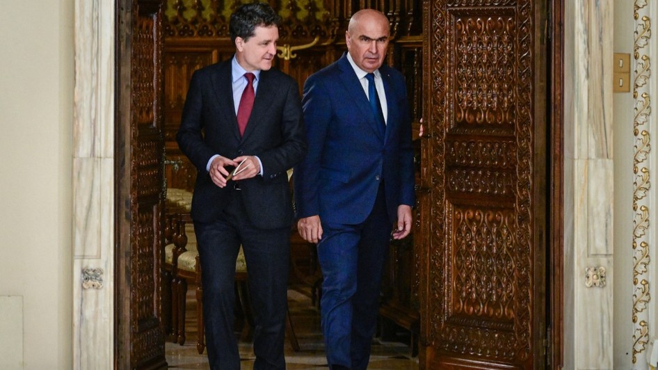 Nicușor Dan și Ilie Bolojan, ambii îmbrăcați în costume elegante, ieșind împreună dintr-o încăpere printr-o ușă masivă din lemn sculptat.