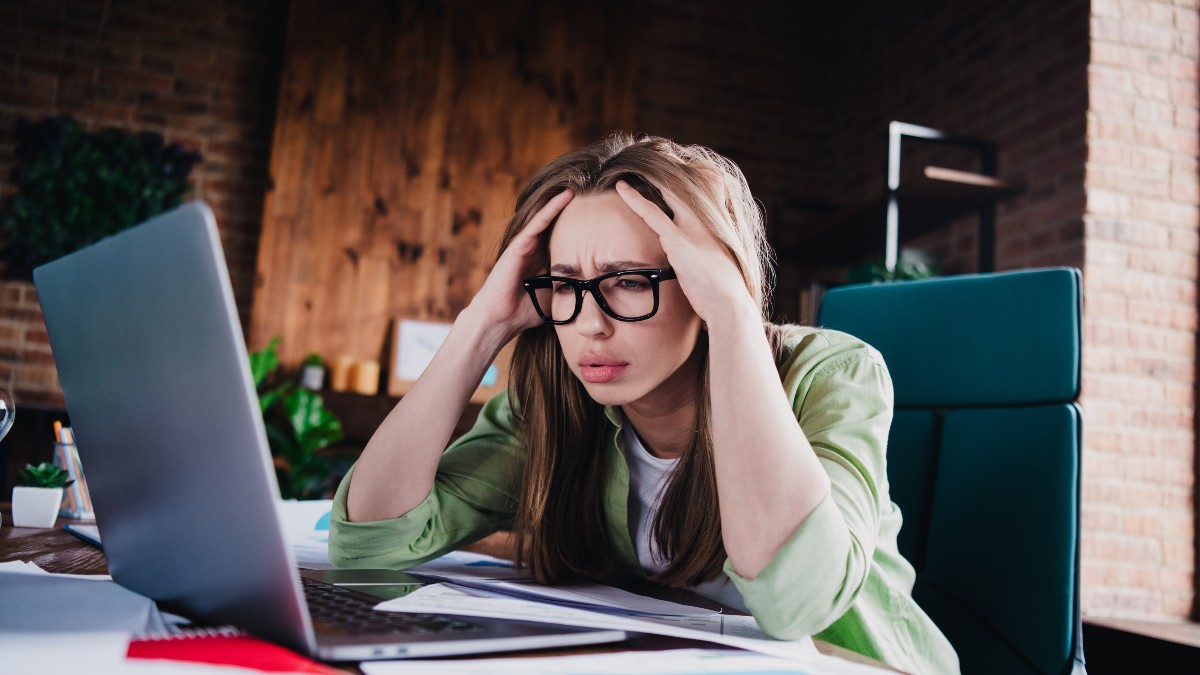 Imaginea o prezintă pe o femeie tânără, cu ochelari, care stă la un birou și se uită la un laptop, având ambele mâini la cap, într-un gest de concentrare intensă sau de stres. Femeia pare să lucreze într-un spațiu modern, cu pereți din cărămidă și lemn, înconjurată de documente și grafice împrăștiate pe birou.