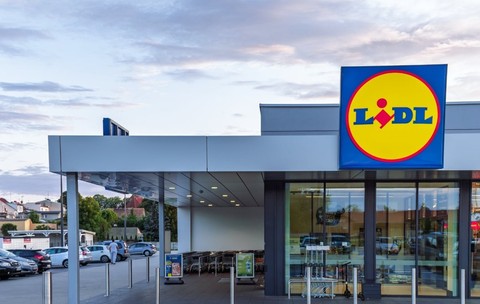 Un magazin Lidl modern este surprins din exterior, cu intrarea largă și logo-ul vizibil deasupra ușilor automate. În fața clădirii se află parcarea și zona pentru cărucioare, sugerând acces facil pentru clienți.
