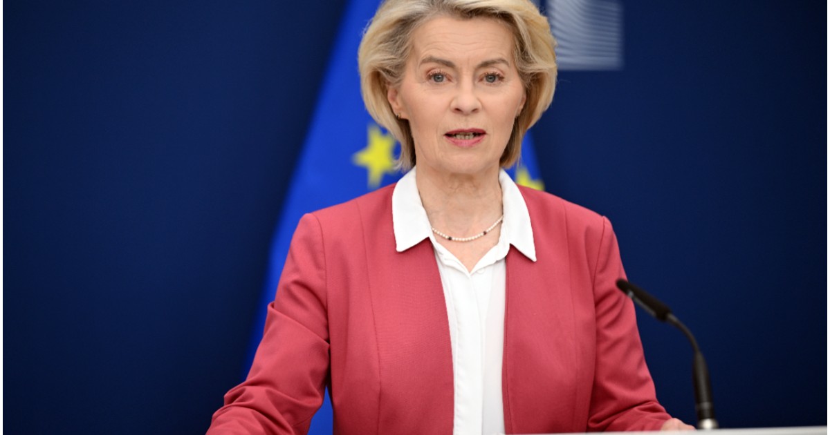 Ursula von der Leyen, îmbrăcată într-un sacou roșu și cămașă albă, în timpul unui discurs, având în spate steagul european