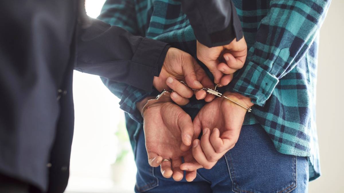 Cadru strans cu mainile unui polistist in timp ce incatuseaza un barbat cu o camasa in carouri verde cu negru si pantaloni de blugi