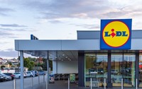 Un magazin Lidl modern este surprins din exterior, cu intrarea largă și logo-ul vizibil deasupra ușilor automate. În fața clădirii se află parcarea și zona pentru cărucioare, sugerând acces facil pentru clienți.