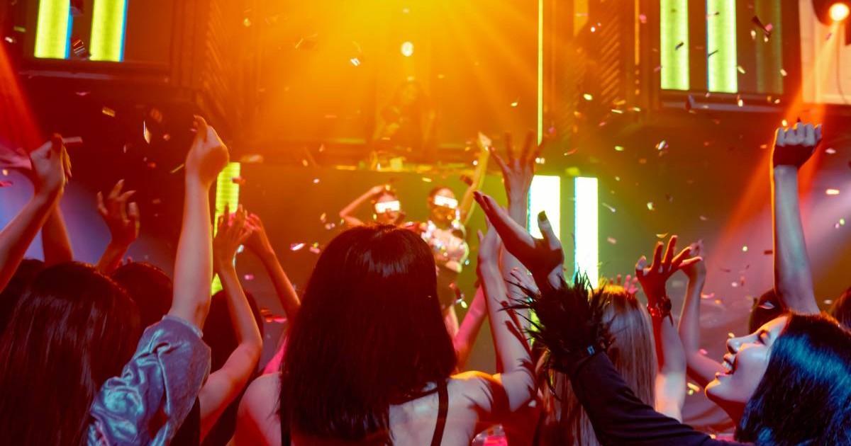 Un grup de femei în timp ce se distrează într-un club din București, laserele luminează încăpere și DJ-ul se vede că pune muzică la pupitru