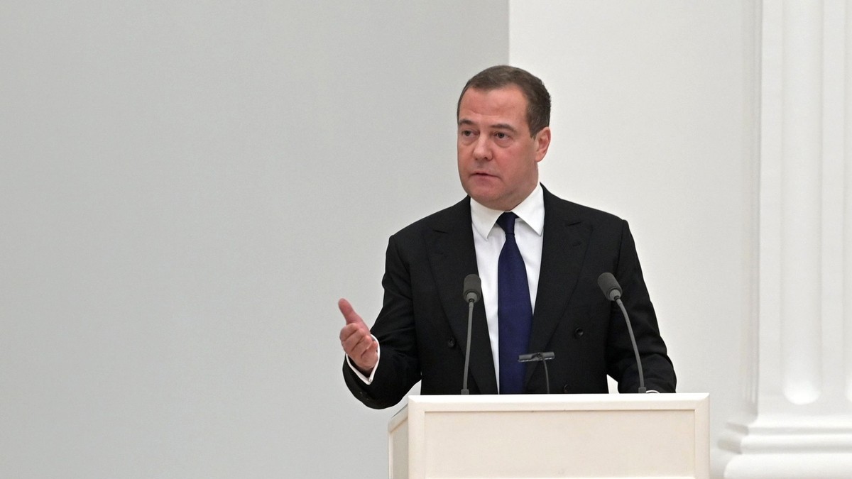 Dmitri Medvedev surprins în timp ce vorbește la microfon, în februarie 2022. Poartă un sacou negru, o cămașă albă și o cravată albastră și ține o mână ridicată