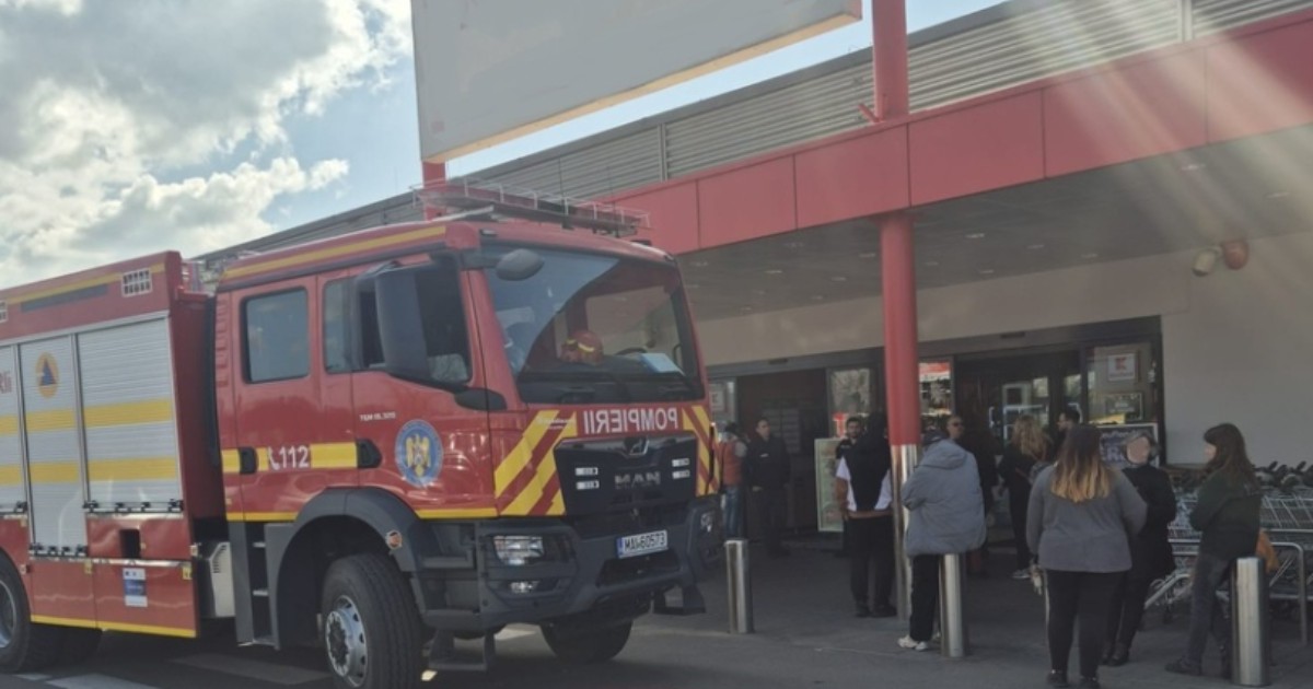 În imagine este ilustrată o mașină de pompieri. În apropiere se află mai mulți oameni, în fața unui supermarket.