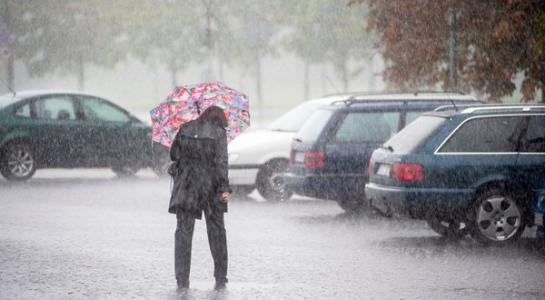 Imaginea prezintă o persoană îmbrăcată în haine închise la culoare care merge sub o umbrelă colorată în timpul unei ploi torențiale, într-o zonă de parcare cu autoturisme.   