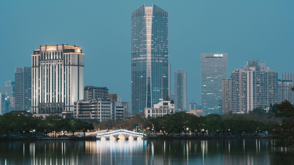 Imaginea prezintă un oraș modern la amurg, cu zgârie-nori iluminați care se reflectă într-un lac liniștit. Un pod arcuit, luminat, traversează apa și accentuează atmosfera calmă și urbană a peisajului.