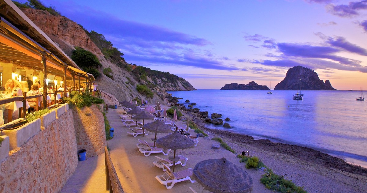 O plajă din Ibiza, Spania, pe înserate. În stânga este o parte dintr-o terasă cu luminile aprinse, iar jos se pot vedea mai multe șezlonguri cu umbrele, iar în dreapta este apă.