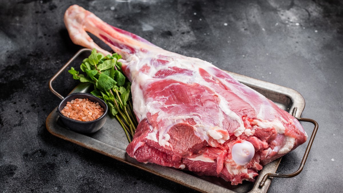 O bucată de carne de miel crudă este așezată pe o tavă metalică, alături de ierburi proaspete și sare grunjoasă. Aspectul sugerează că ingredientele sunt pregătite pentru gătit, într-un cadru simplu și rustic.