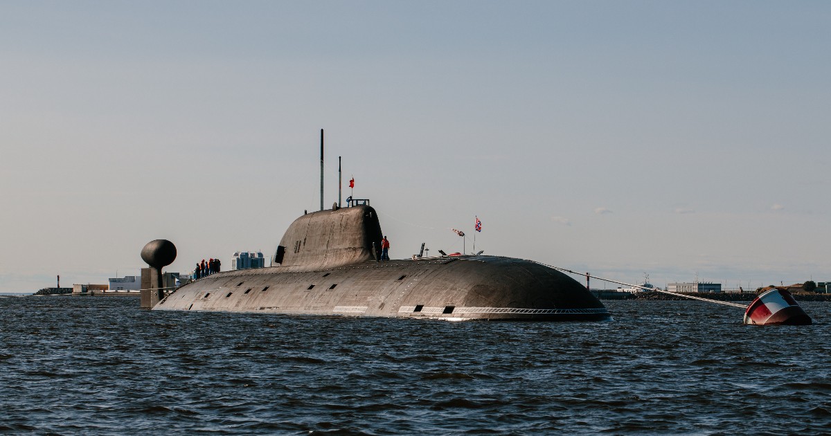 Un submarin mare, de culoare închisă și cu o siluetă masivă, plutește pe suprafața unei ape agitate sub un cer senin, afișând pavilionul național la catarg. O porțiune vizibilă a cocii sale iese din apă, iar câțiva oameni pot fi zăriți pe puntea sa, sugerând că este andocat sau se deplasează în apropierea coastei.