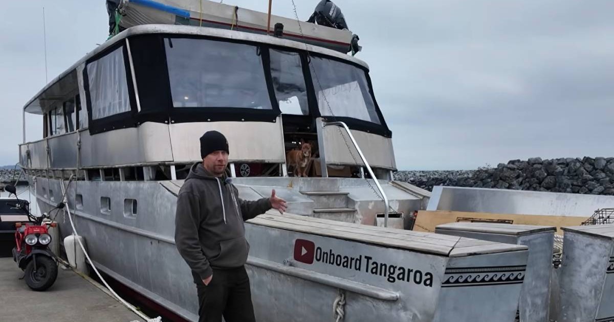 Un barbat, cu caciula pe cap, hanorac gri si pantalon negru, in marina, arata spre iahtul Tangarao, barca pe care a renovat-o cu sotia sa