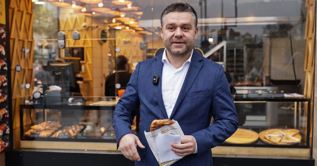 Ciprian Ciucu, cu un sacou albastru și cămașă albă, ține un covrig în mână, în fața unei covrigării din Bucuresti - 13 Noiembrie 2025