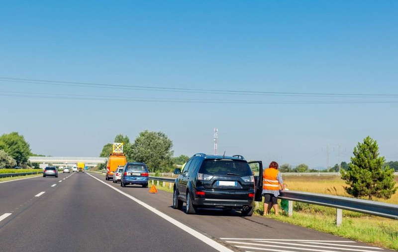 Mai multe mașini oprite pe banda de urgență de pe autostradă. Prima mașină este o mașină neagră, iar în dreapta ei este o femeie cu vestă reflectorizantă. În fața mașinii negre sunt alte două mașini oprite, care au triunghiul reflectorizant montat.