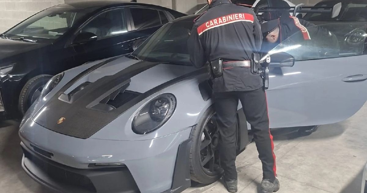 Imaginea surprinde doi carabinieri lângă un Porsche 911 elegant, de culoare gri mat. Scena pare să se desfășoare într-un garaj, unde se află mai multe mașini.