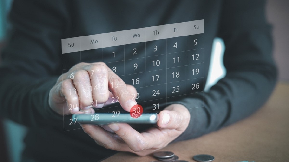 Ce este regula de 30 de zile și cum te ajută să economisești - Persoană care programează o dată pe telefon, cu bancnote și monede pe masă Sursa foto: Shutterstock