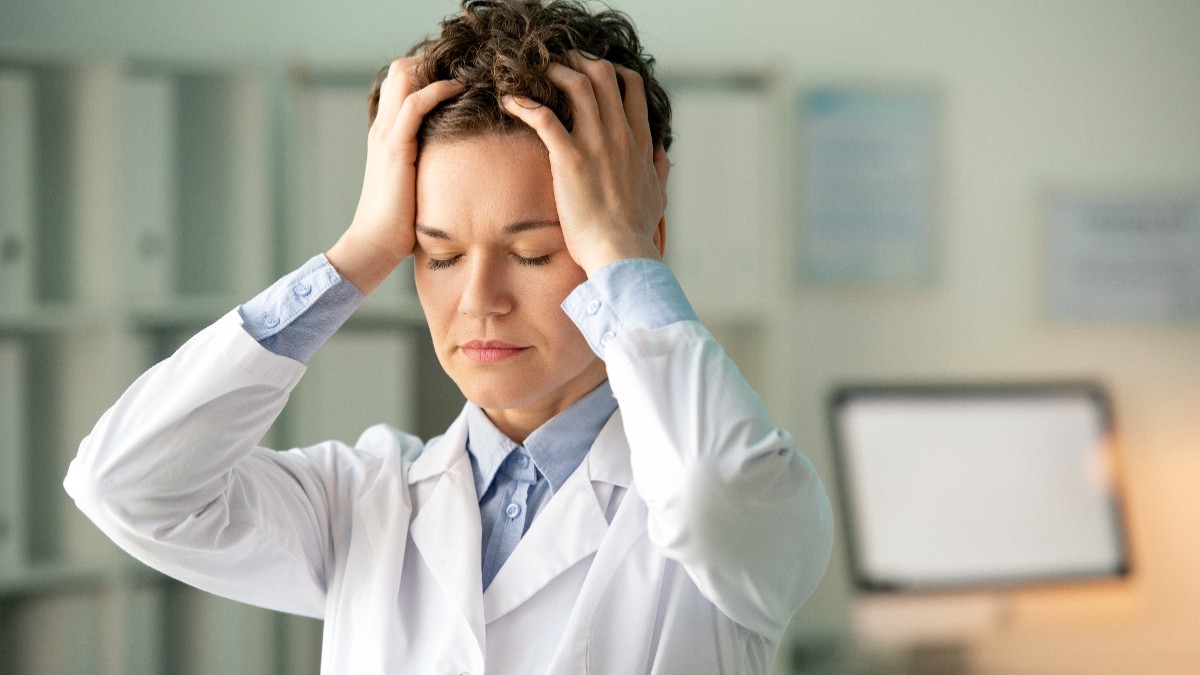 O farmacistă care își ține mâinile pe cap, pare epuizată și dezamăgită. Femeia poartă halat alb