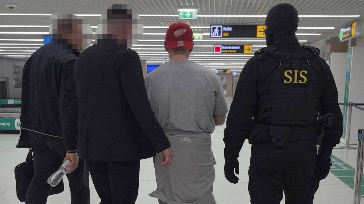 Un barbat in haine gri si o sapca rosie pe cao, inconjurat de mai multi agenti SIS in negru