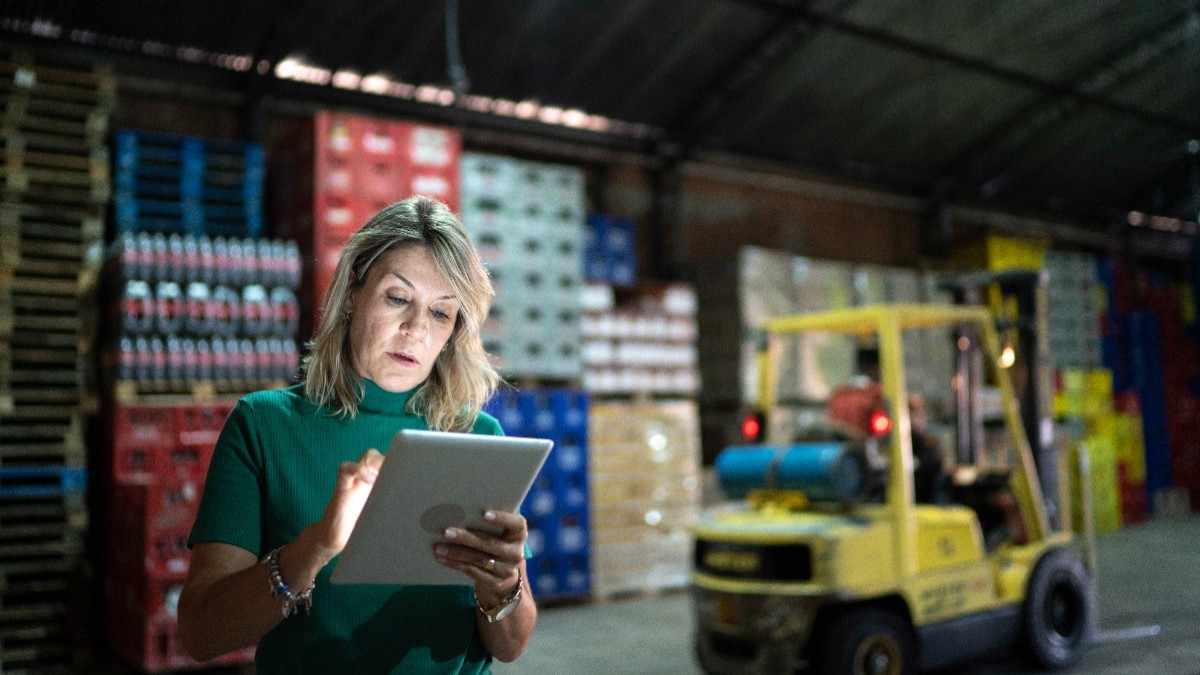 O femeie îmbrăcată într-o bluză verde, utilizând o tabletă digitală într-un depozit industrial cu mărfuri stivuite și un motostivuitor galben în fundal.