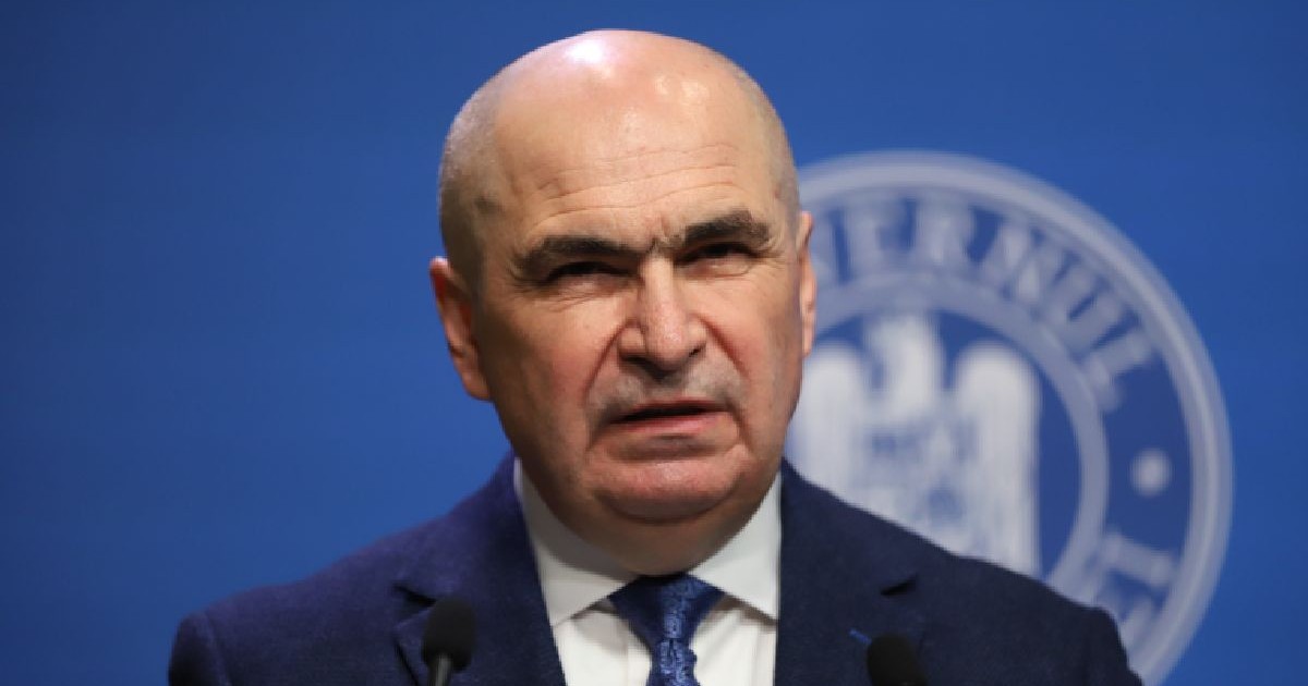 Imaginea îl prezintă pe Ilie Bolojan, îmbrăcat într-un costum bleumarin și cămașă albă, vorbind în fața unor microfoane pe fundalul unui panou oficial albastru ce conține o stemă.