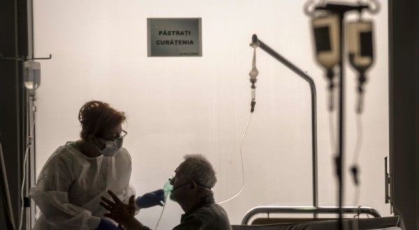 pacient stand pe pat intr-un salon de spital cu o perfuzie la capul patului. Langa el sta o femeie care-i pune masca de oxigen pe gura