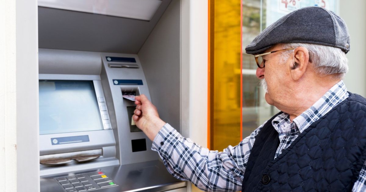 Bătrân îmbrăcat cu cămașă și vestă de culoare albastră, cu bască pe cap, introduce un card într-un bancomat