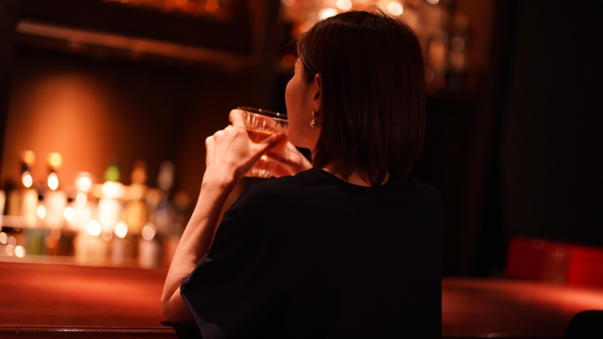 În imagine este ilustrată o femeie care ține un pahar cu alcool în mână, în timp ce stă la un bar