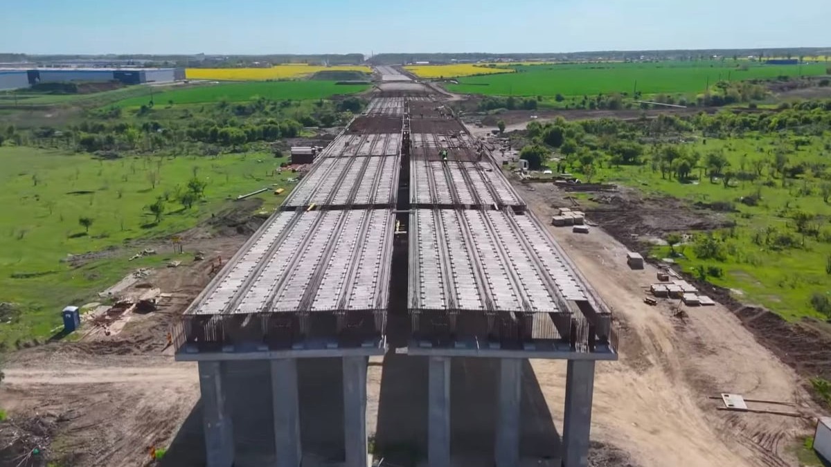 Vedere aeriană de-a lungul unui viaduct aflat în construcție pentru o autostradă, cu grinzi masive de beton sprijinite pe piloni, traversând o zonă de câmpie cu vegetație și terenuri agricole.