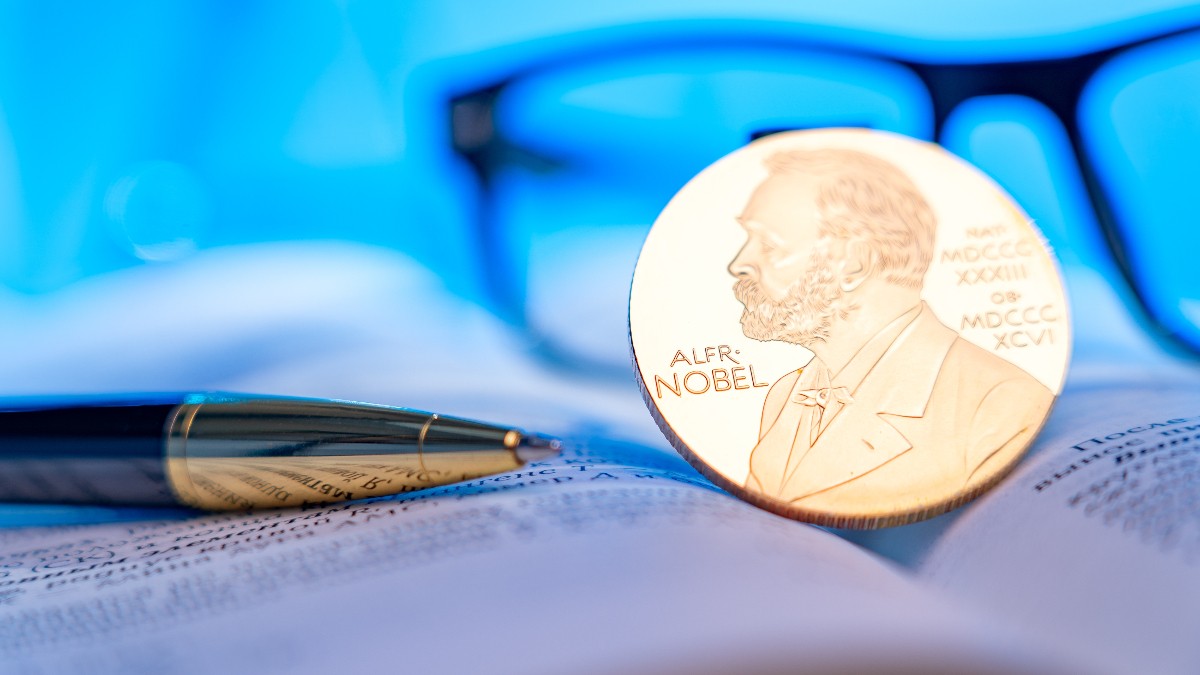Imaginea este un prim-plan cu focalizare superficială care surprinde o replică a medaliei Nobel de culoare aurie, sprijinită pe o carte deschisă, având gravat portretul lui Alfred Nobel. Pe fundalul estompat de culoare albastră se văd o carte deschisă cu text lizibil, ochelari și un instrument de scris elegant, sugerând un mediu academic sau de cercetare.