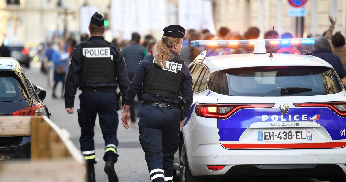 Cei doi polițiști francezi (un bărbat și o femeie), îmbrăcați în uniforme închise cu veste tactice inscripționate "POLICE", se îndepărtează de o mașină de patrulă albă cu girofarurile aprinse. Ofițerii patrulează pe o stradă urbană, cu o mulțime de oameni neclară în fundal, sugerând prezența lor în timpul unui eveniment public.