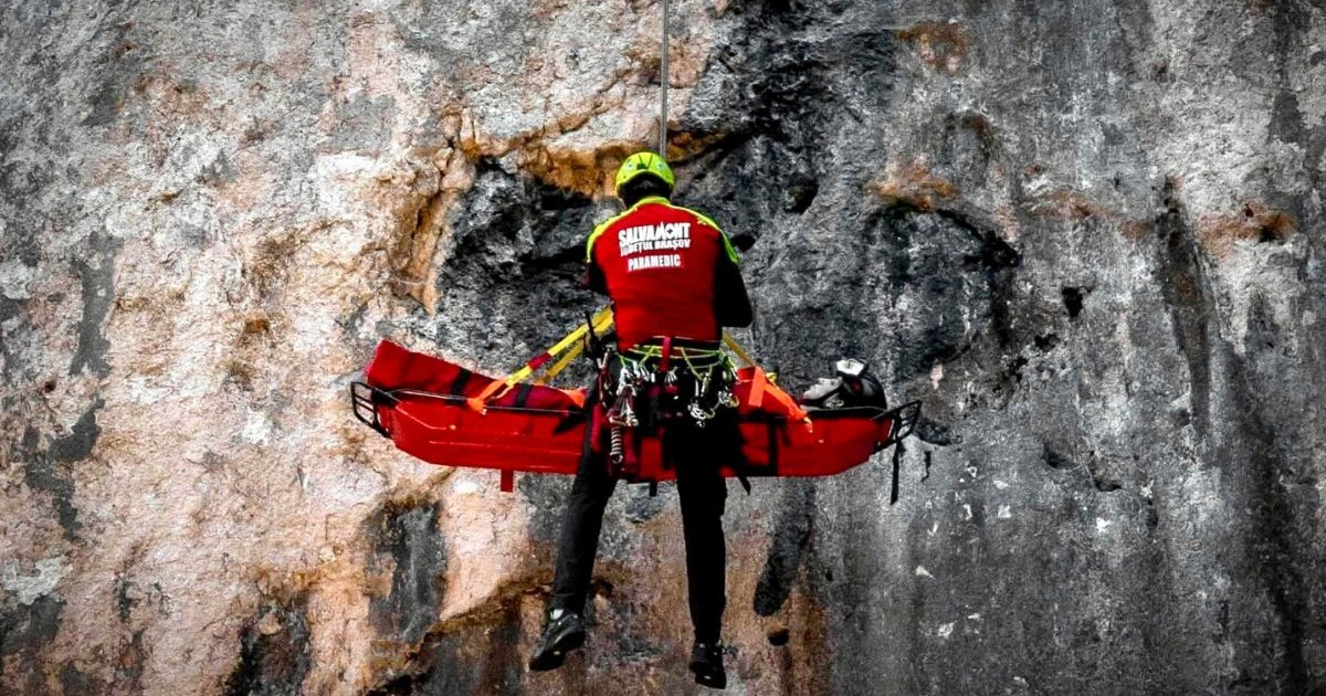 o persoană văzută din spate, îmbrăcată într-o vestă roșie și cască galbenă, suspendată cu ajutorul unor corzi în fața unei stânci verticale. Persoana stă lângă o targă rigidă roșie, prinsă de sistemul de prindere al corzilor. În fundal se vede suprafața aspră, gri și brună a stâncii. Pe vesta persoanei sunt inscripții albe cu textul „Salvamont Județul Brașov - Paramedic”