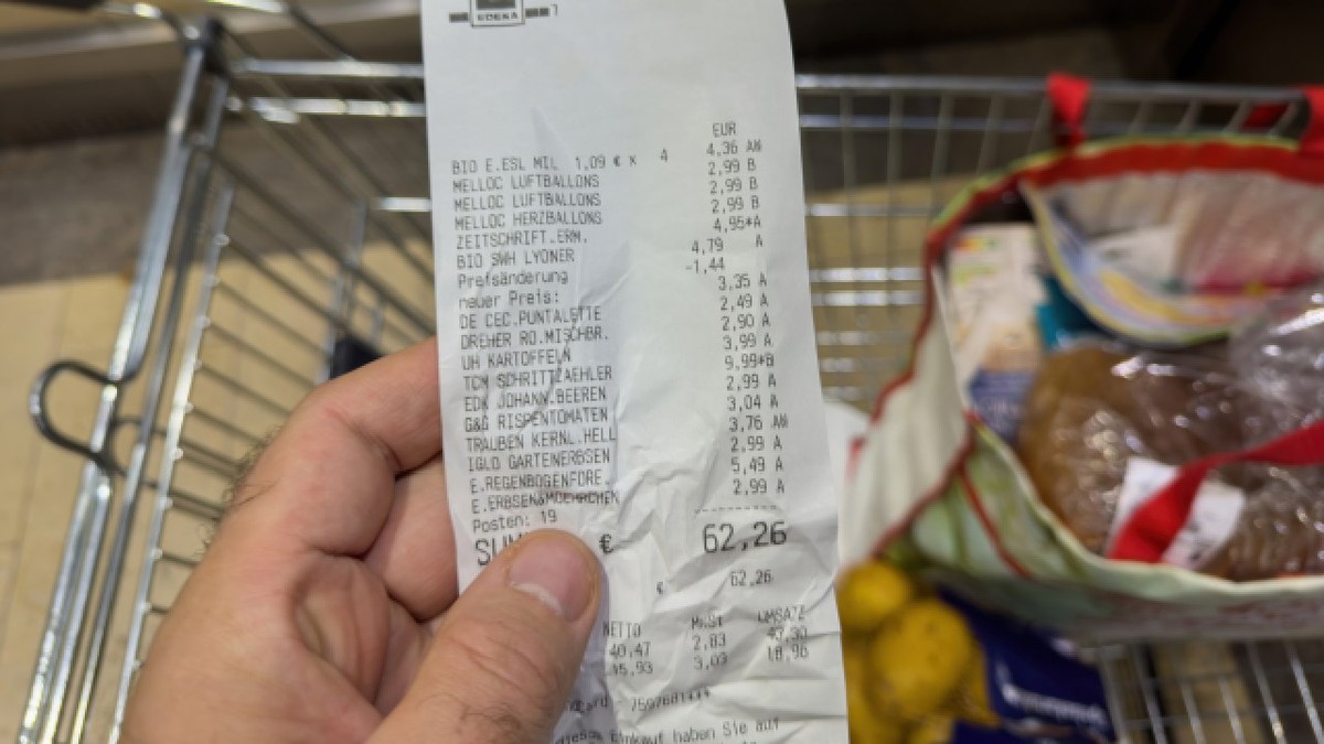 Frankfurt, Germania - 24 octombrie 2023: Mână de bărbat ține oun bon în valoare totală de 62,26 euro la supermarketul german Edeka, cu un cărucior de cumpărături și o pungă reutilizabilă plină în fundal, simbolizând inflația.