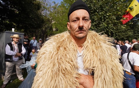 Un bărbat purtând o căciulă neagră și un cojoc din blană de oaie, privind fix spre cameră în mijlocul altor participanți la un protest în aer liber, unde se observă și un steag al României.