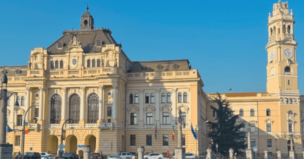 O clădire în nuanțe de galben pal/bej deschis. În partea dreaptă a clădirii este un turn înalt cu ceas