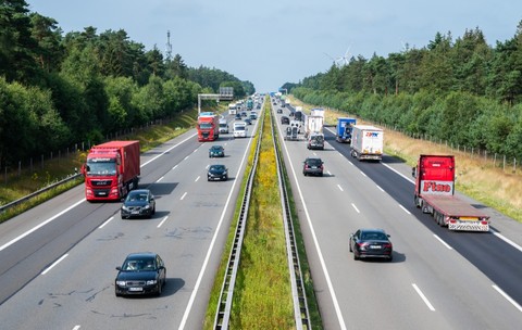 Imaginea prezintă o privire de sus asupra unei autostrăzi aglomerate, pe care circulă numeroase camioane și autoturisme în ambele sensuri, traversând o zonă împădurită.