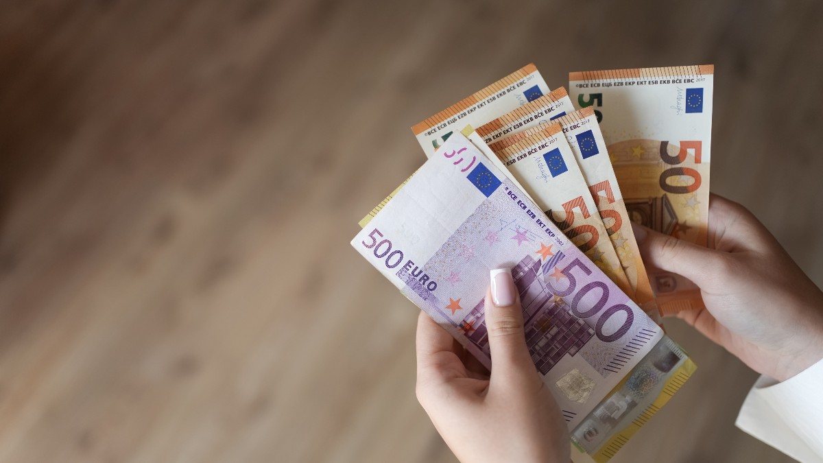 Imaginea surprinde un prim-plan al unor mâini care țin și numără mai multe bancnote Euro, inclusiv o bancnotă violet de 500 de Euro și bancnote portocalii de 50 de Euro. Mâinile au o manichiură îngrijită, iar fundalul este estompat și arată o suprafață de lemn.