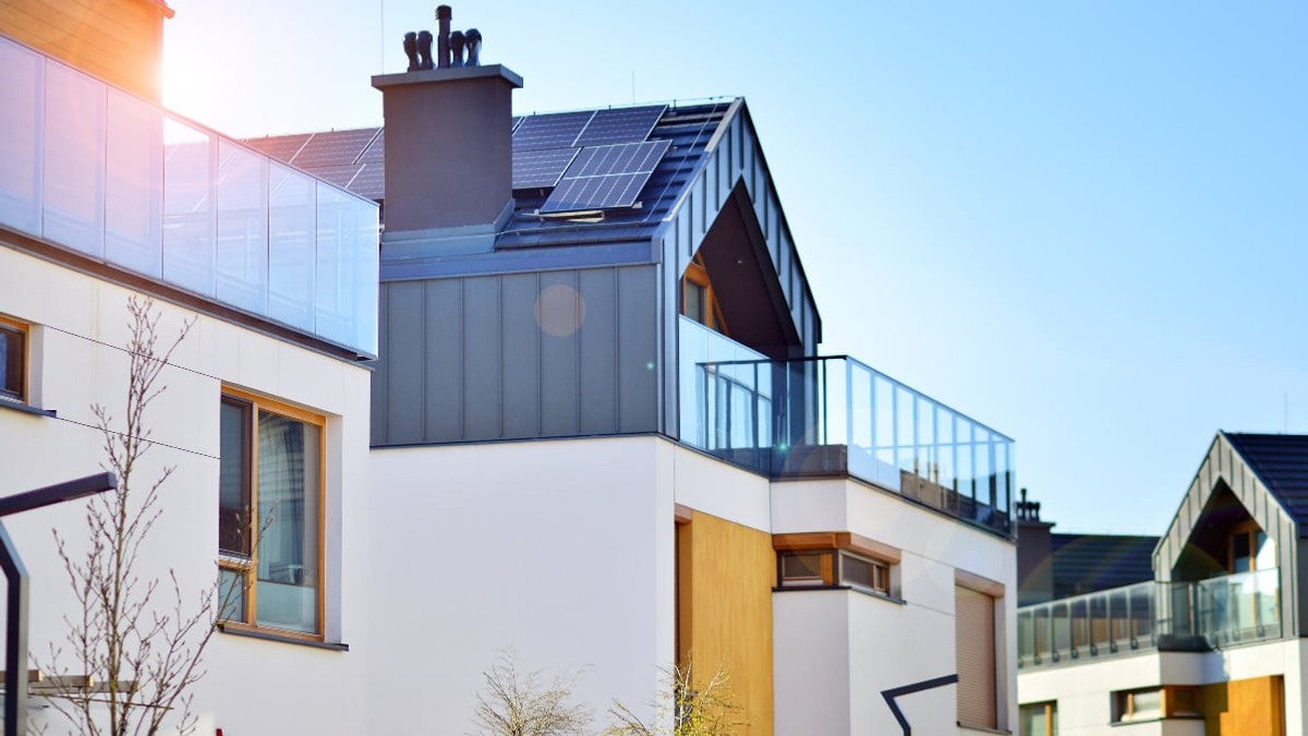Imagini prezintă case moderne cu design arhitectural minimalist, având balcoane cu balustrade din sticlă și un horn mare de culoare închisă. Pe acoperișul înclinat al clădirii principale sunt montate panouri solare, totul fiind scăldat în lumina caldă a soarelui sub un cer senin.