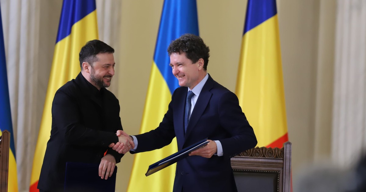 Volodimir Zelenski, îmbrăcat într-o ținută neagră de tip casual-militar, dă mâna zâmbind cu Nicușor Dan, care poartă un costum bleumarin formal cu cravată și ține un dosar cu documente în mână. Cei doi lideri se află în fața unui rând de steaguri ale României și Ucrainei, marcând un moment oficial de cooperare într-un cadru instituțional solemn.